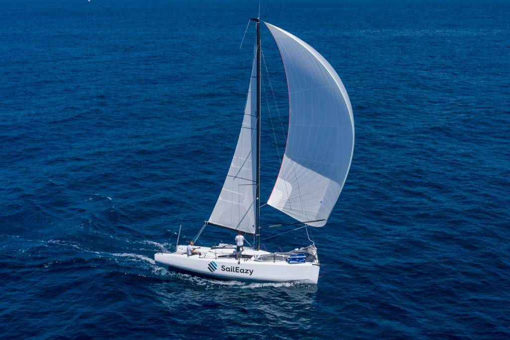 Voilier SailEazy, MMW33 qui se nomme Cipango et qui est basé à Marseille. Il est de profil, naviguant vers la droite avec une grand voile blanche, un Spinaker blanc et une coque bloanche floqué SailEazy.