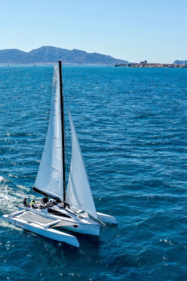 SailEazy, voilier en libre-service. Le Trimaran, tricat 30 qui se nomme breva navigue vers le bas droit de l'écran. Il est vu de haut en contre plongé. Il a une grand voile blanche, un spinnaker blanc et les flotteur déplié sont aussi blanc avec saileazy floqué dessus. La mer est bleu foncé et assez calme.