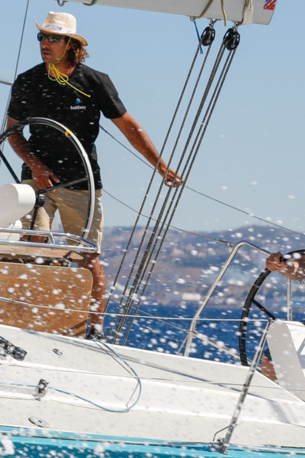 SailEazy, location voilier en lobre-service. On voit un équipage composé de 6 personnes sur Ty Punch, un RM 1070. Un coach est devant la barre à roue tribord et un équipier sur celle babord. Il y a des éclaboussures.