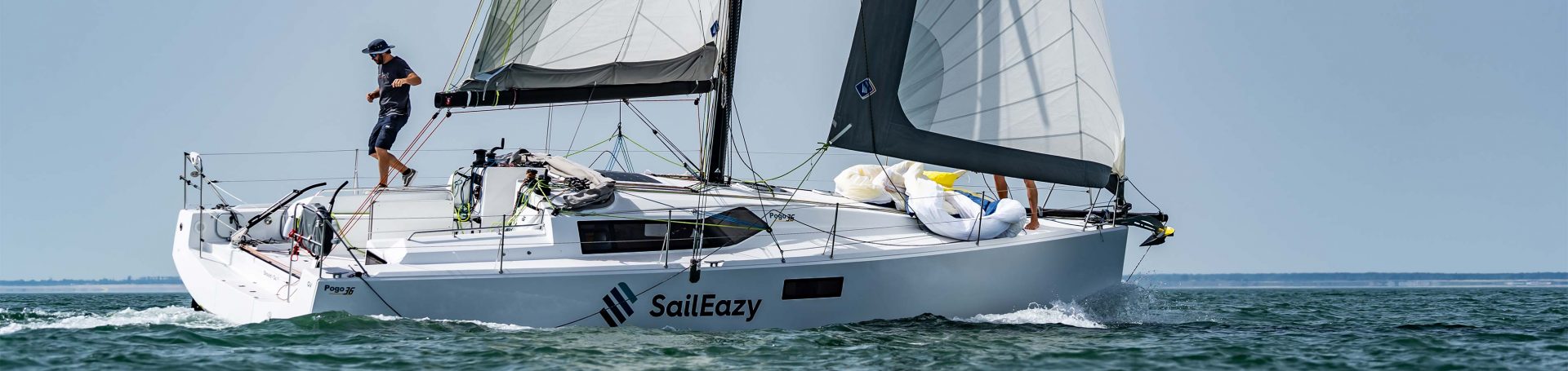 SailEazy, location voilier en lobre-service. On voit un équipage composé de 6 personnes sur Ty Punch, un RM 1070. Un coach est devant la barre à roue tribord et un équipier sur celle babord. Il y a des éclaboussures.