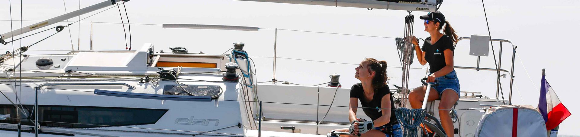 SailEazy, voilier en libre-service. Nous voyons deux femme vetu d'un t-shirt noir avec le logo de saileazy, une à la barre, l'autre au winch. Elles navigues sur un Elan E3. Elles soont souriante. 
