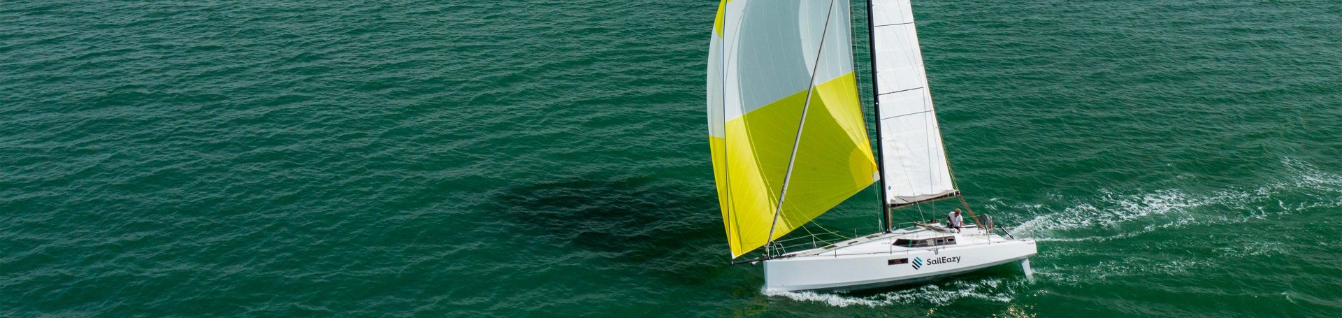 SailEazy, voilier en libre-service. C'est un voilier, modèle Pogo 36 qui se nomme Should I Go. Il navigue vers la gauche. Il est de profile. Il a une grand voile blanche, un spinnaker jaune et blanc et une coque blanche floqué SailEazy. La mer a une teinte un peu verte. 